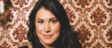 A woman with black hair smiles at the camera. She wears a black blouse. Behind her, a patterned wallpaper with a brown and beige design is visible. An orange lamp is mounted on the wall to the left.