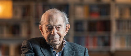 Ein älterer Mann mit weißem Haar, der einen grauen Anzug und ein gestreiftes Hemd trägt, lächelt und lehnt sich an einen Glastisch. Dahinter steht ein mit Büchern gefülltes Bücherregal.