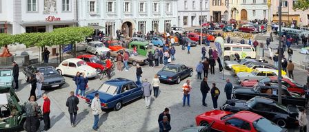 A bustling town square filled with various vintage cars parked in rows, surrounded by buildings and people walking around.