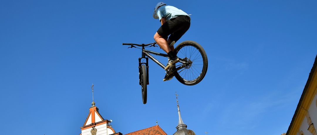 Eine Person führt einen Fahrradtrick in der Luft über einem Gebäude mit dem Schild 'Balhaus' und einem Kirchturm aus. Der Himmel ist klar und blau.