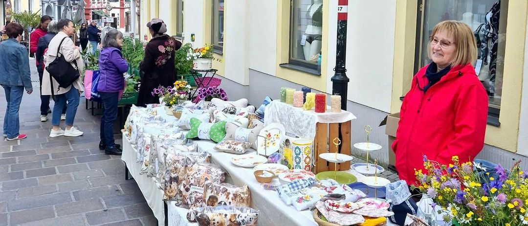 Menschen schlendern eine Straße entlang, an deren Seiten sich Geschäfte befinden. Eine Frau in einer roten Jacke steht neben einem Tisch mit verschiedenen Artikeln, darunter Blumen und Kunsthandwerk.