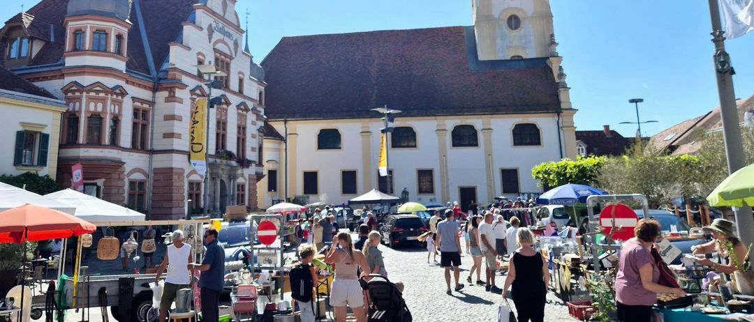 Ein sonniger Tag auf einem Stadtplatz mit einem geschäftigen Flohmarkt. Im Hintergrund steht ein großes Kirchengebäude. Menschen gehen herum und stöbern in Ständen.
