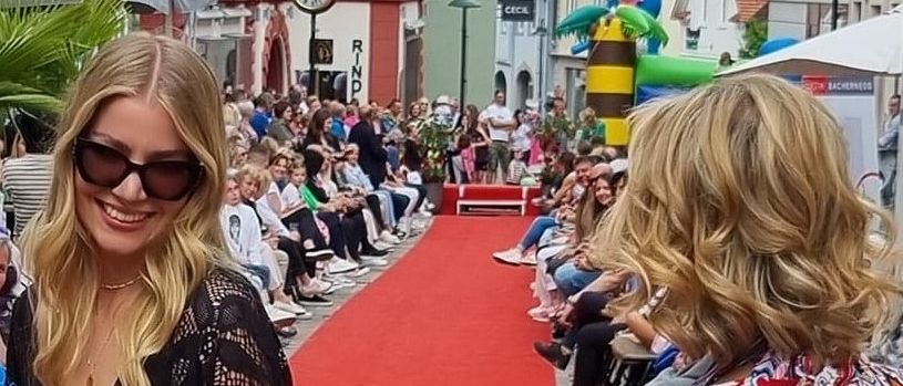 A woman with blonde hair wearing sunglasses and a black outfit stands on a red carpet. Behind her, several people sit on chairs on the side. An inflatable is in the background.