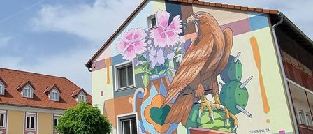 Ein Haus mit einem großen Wandbild eines Vogels mit Blumen und Früchten darauf. Es hat ein rotes Dach und einen Balkon mit Geländern. Ein weiteres Haus ist im Hintergrund.