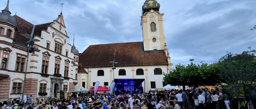 Eine lebhafte Menge genießt Sitzgelegenheiten vor einer historischen Kirche, unter einem teilweise bewölkten Himmel. Tische sind zum Essen gedeckt, mit einer Bühne im Hintergrund.