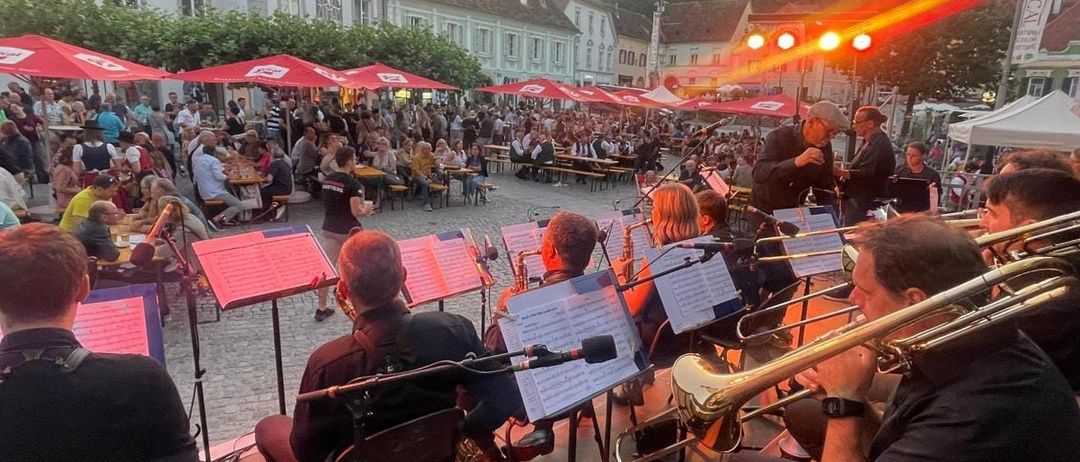 Eine Band spielt auf einer Bühne unter roten Regenschirmen vor einem Publikum. Mehrere Menschen sitzen auf Stühlen, während andere im Hintergrund stehen. Gebäude und Bäume sind in der Ferne zu sehen.