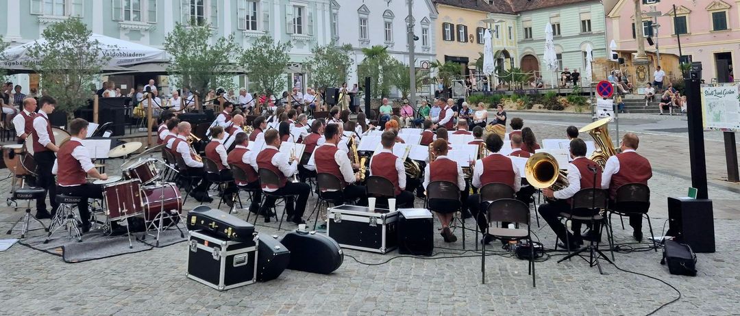 Ein Open-Air-Konzert mit Blechblasinstrumenten auf einem Stadtplatz mit klassischen Gebäuden und einem bewölkten Himmel.