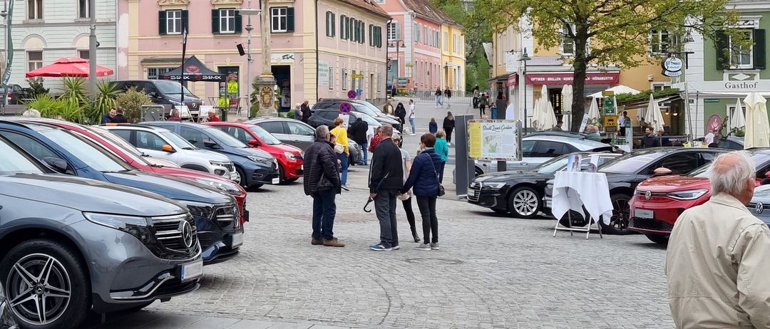 A town square with cobblestone pavement, trees, and parked cars. People walk and stand around. Buildings with different colors and designs line the square.