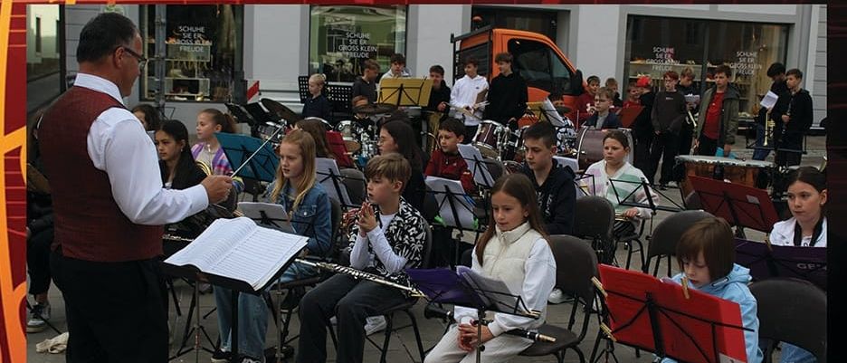 Ein Sommerkonzert mit einer Band von Kindern und einem Mann, der vor ihnen steht und einen Taktstock hält. Die Kinder spielen verschiedene Instrumente. Das Plakat ist auf Deutsch.