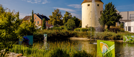 Ein Turm mit einem Brunnen vor einem Teich. Ein Haus und Bäume im Hintergrund.