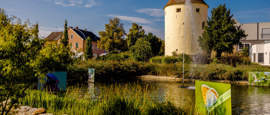 Ein Turm mit einem Brunnen vor einem Teich. Ein Haus und Bäume im Hintergrund.