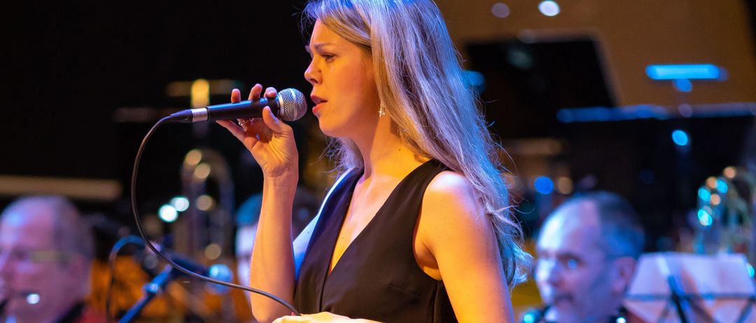 Eine Frau singt in ein Mikrofon während einer Bühnenaufführung mit einer Band. Sie trägt ein schwarzes Kleid und hält ein Mikrofon. Hinter ihr spielen Musiker verschiedene Instrumente in einem schwach beleuchteten Raum.