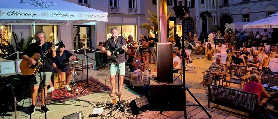 Ein Abendkonzert einer Musikband vor einem Gebäude mit einem sitzenden Publikum. Die Bühne hat Lautsprecher, Mikrofone und einen Teppich. Das Publikum genießt Getränke und der Himmel wird von Straßenlaternen beleuchtet.