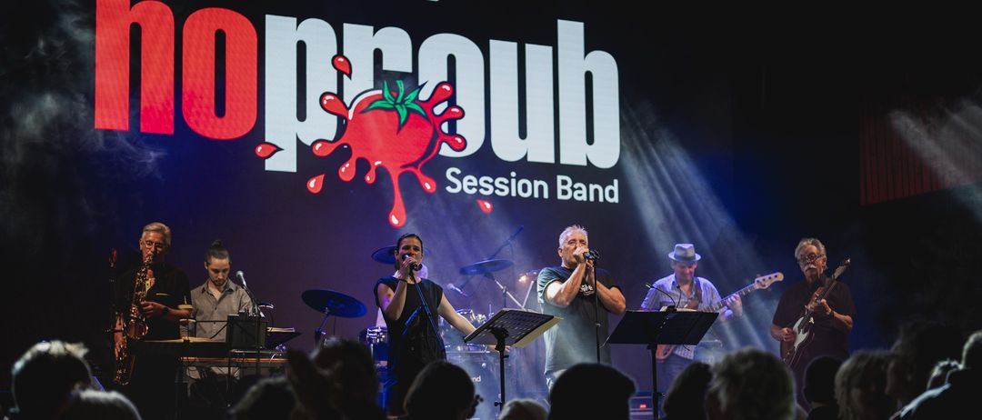 Eine Band tritt vor einem Publikum auf der Bühne auf, mit einem roten Tomaten-Spritzlogo.