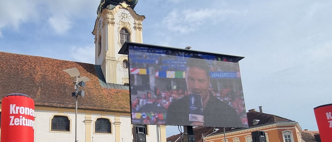 Eine große Außenleinwand zeigt einen sprechenden Mann vor einem turmartigen Gebäude bei teilweise bewölktem Himmel.