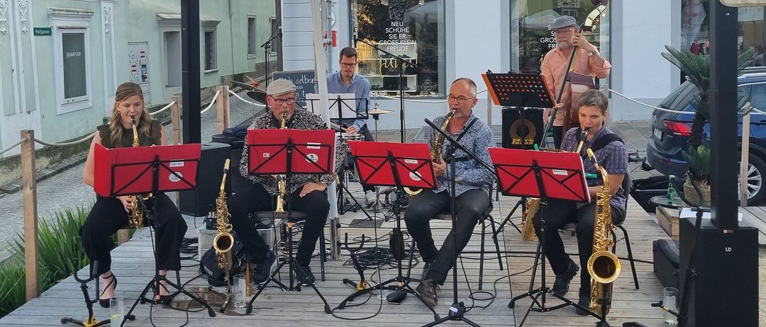 Eine Gruppe Musiker spielt auf einer Holzplattform unter einem großen Regenschirm vor einem Gebäude. Sie spielen Saxophone und andere Instrumente.