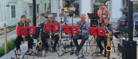 Eine Gruppe Musiker spielt auf einer Holzplattform unter einem großen Regenschirm vor einem Gebäude. Sie spielen Saxophone und andere Instrumente.
