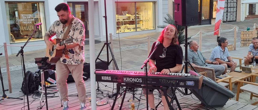 Zwei Musiker spielen auf einer Holzterrasse mit einem Korg Kronos Keyboard, rosa Beleuchtung und einem Mikrofon. Ein Mann spielt Gitarre unter einem Schirm.