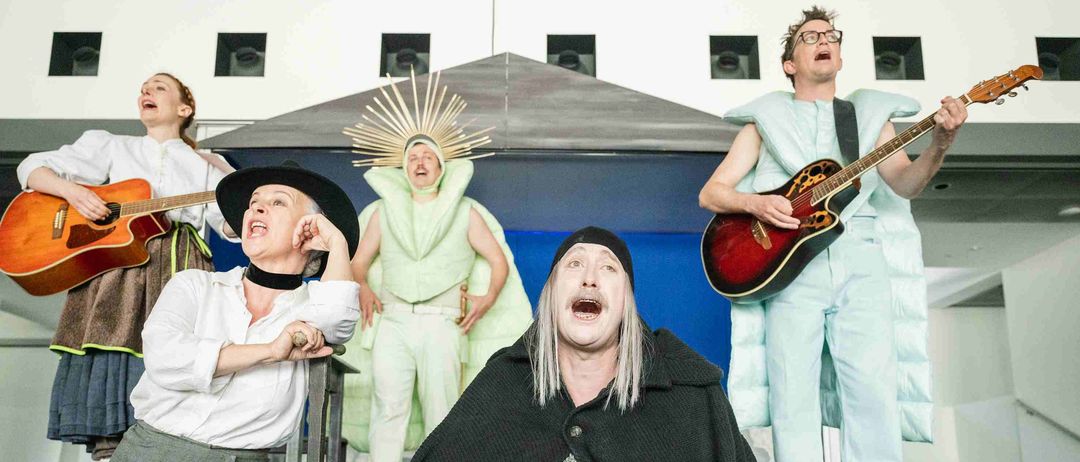Eine Gruppe von Darstellern auf einer Bühne, eine Person sitzt und schreit, während andere stehen und Musikinstrumente spielen.
