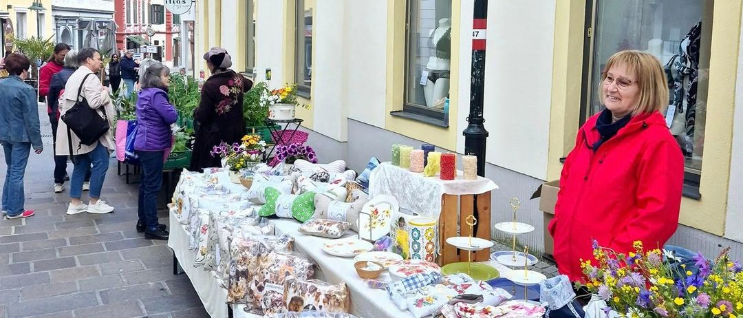 Ein Straßenmarkt mit Leuten, die Gegenstände wie Handwerkskunst, Blumen und Kissen betrachten. Eine Frau in einer roten Jacke steht hinter dem Tisch.