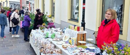 Ein Straßenmarkt mit Leuten, die Gegenstände wie Handwerkskunst, Blumen und Kissen betrachten. Eine Frau in einer roten Jacke steht hinter dem Tisch.