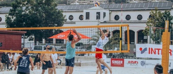 Eine Gruppe von Menschen spielt Beachvolleyball auf einem sandigen Spielfeld mit einem Netz, Gebäuden und Bäumen im Hintergrund. Ein Mann springt und schlägt den Ball.