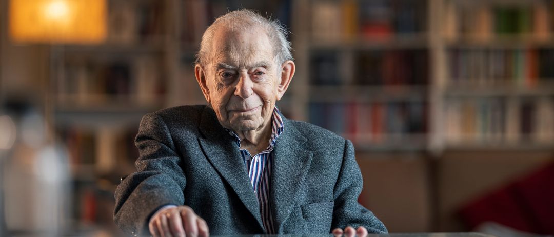 Ein älterer Mann lächelt, sitzt an einem Glastisch mit Bücherregalen im Hintergrund.
