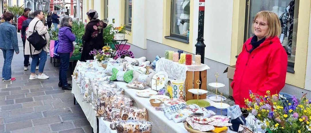 Eine Frau steht hinter einem Tisch voller handgefertigter Waren. Viele Menschen gehen vor dem Tisch. Das Gebäude hat gelbe Verkleidungen und Glasfenster.
