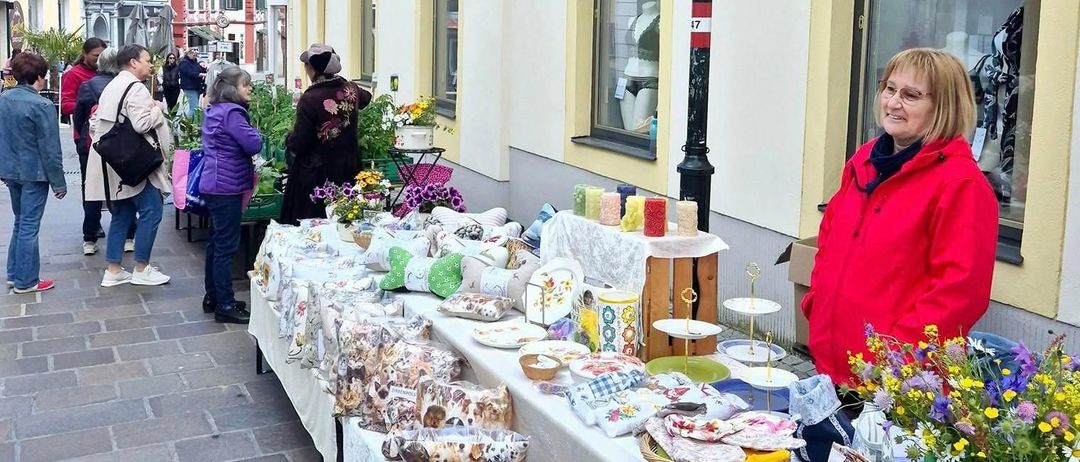 Eine Frau in einem roten Mantel steht neben einem Tisch mit verschiedenen Artikeln zum Verkauf. Der Tisch ist mit einem weißen Tuch bedeckt und mit farbenfrohen Artikeln gefüllt, darunter Blumen, Süßigkeiten und andere Waren. Hinter ihr gehen Menschen auf der Kopfsteinpflasterstraße, und Gebäude mit gelben Verzierungen sind sichtbar.