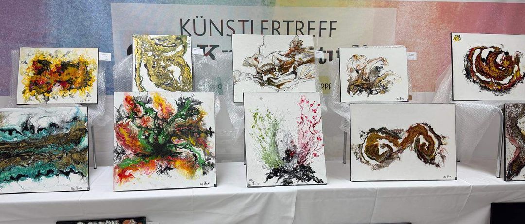 Eine Ausstellung bunter abstrakter Gemälde, die auf einem weißen Tisch angeordnet sind. Die Gemälde haben verschiedene Größen und Stile. Der Hintergrund trägt den Schriftzug Kunstlertreff auf Deutsch.