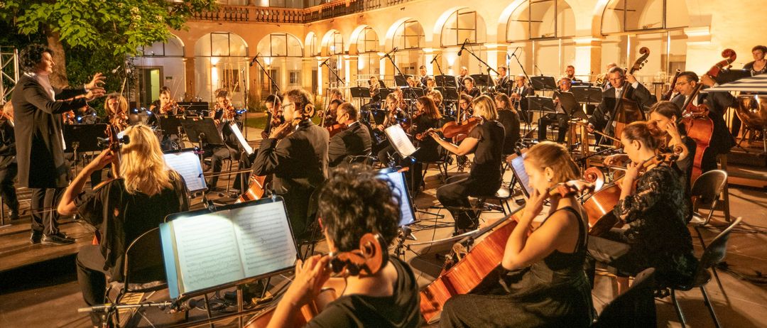 Ein Orchester spielt ein Musikstück in einem Innenhof eines historischen Gebäudes mit Rundbogenfenstern. Die Musiker spielen Violinen, Celli und Trompeten.