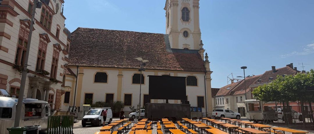 Eine Reihe von Holzbänken steht vor einer Kirche mit einem hohen Turm und einer Uhr. Es sind Fahrzeuge vor der Kirche geparkt.
