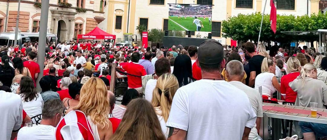 Eine Menschenmenge versammelt sich vor einem historischen Gebäude mit Glockenturm. Viele tragen rote Shirts, und eine große Leinwand zeigt ein Fußballspiel.