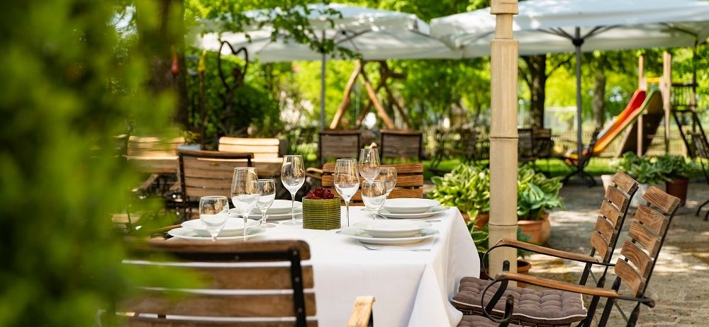 Ein Outdoor-Esstisch mit Tellern, Weingläsern und einer Obstschale. Der Tisch ist von Holzstühlen umgeben. Im Hintergrund befinden sich Bäume, Pflanzen und Sonnenschirme.
