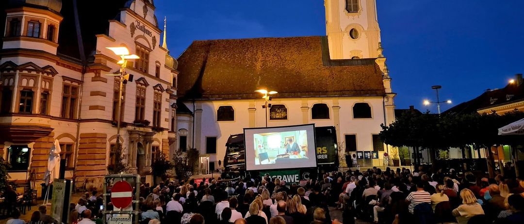 Eine Menschenmenge versammelt sich nachts vor einer Kirche für eine Vorführung, ein Lastwagen projiziert ein Video eines Mannes beim Kochen.