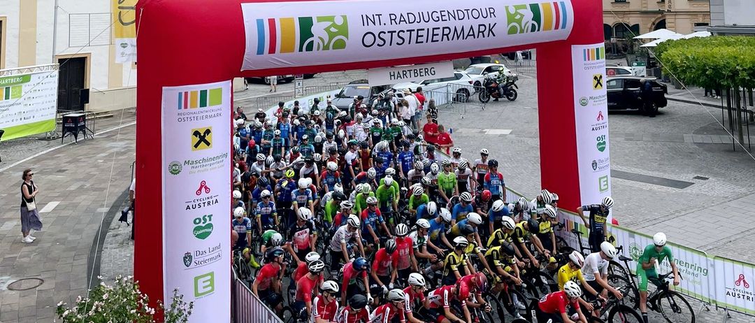 Eine große Gruppe von Radfahrern mit Helmen und Radsporttrikots steht in einer Reihe an der Startlinie eines Rennens vor einem historischen Gebäude.