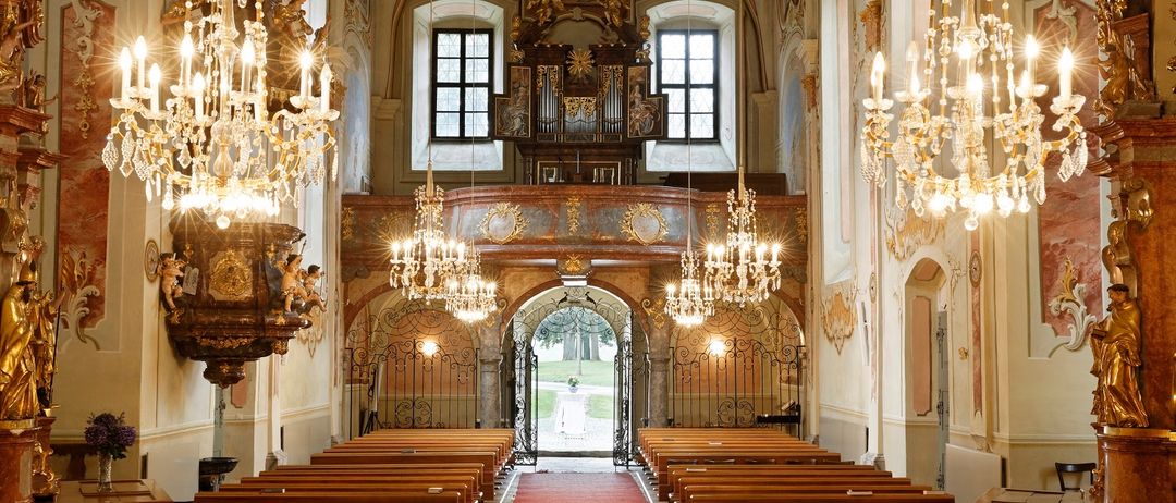 Ein reich verzierter Kircheninnenraum mit hölzernen Kirchenbänken, einem roten Teppich und aufwendigen Kronleuchtern. Die Wände sind mit aufwendigen Designs und Gemälden verziert.