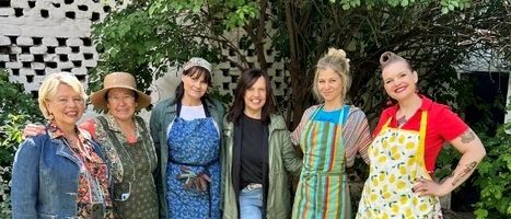 Sechs Frauen in Schürzen stehen in einem Garten und lächeln für ein Foto, mit einem Baum und einem Haus im Hintergrund.
