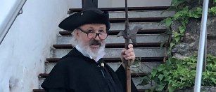 Ein älterer Mann in einem schwarzen Gewand und Hut, der einen Stab hält, steht auf einer Steintreppe mit grünen Kletterpflanzen.