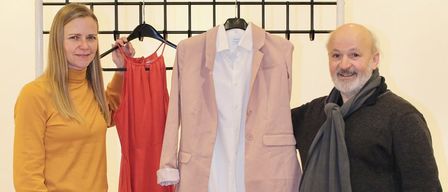 Ein Mann und eine Frau stehen neben einem Tisch mit Kleidern an Kleiderbügeln und gefalteten oben. Die Frau hält ein rotes Kleid und der Mann hält eine rosa Jacke.