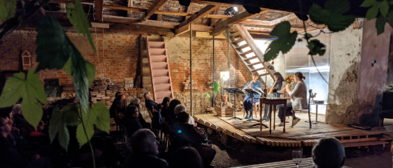 Eine Gruppe von Menschen versammelt sich an einem rustikalen Ort im Freien und sieht sich eine musikalische Aufführung auf der Bühne an. Die Künstler sitzen an einem Tisch mit Musikinstrumenten.