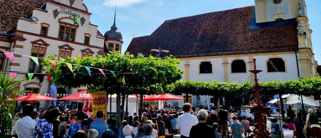 Ein geschäftiger Platz voller Menschen, in der Nähe eines großen Gebäudes mit Glockenturm, unter einem teilweise bewölkten Himmel, mit Bäumen und roten Regenschirmen.