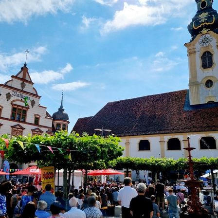 Ein überfüllter Außenbereich mit Menschen, Zelten und Gebäuden, eines mit einem Glockenturm. Bäume und Pflanzen sind vorhanden, ebenso wie Flaggen und Banner.