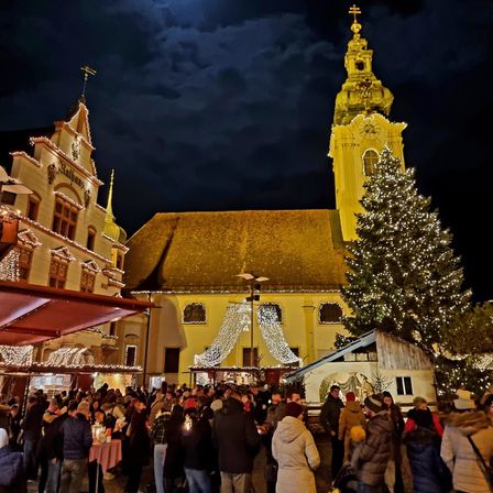 Ein überfüllter Platz vor einer Kirche bei Nacht, geschmückt mit Weihnachtsdekorationen und einem großen Weihnachtsbaum. Der Mond scheint hell.