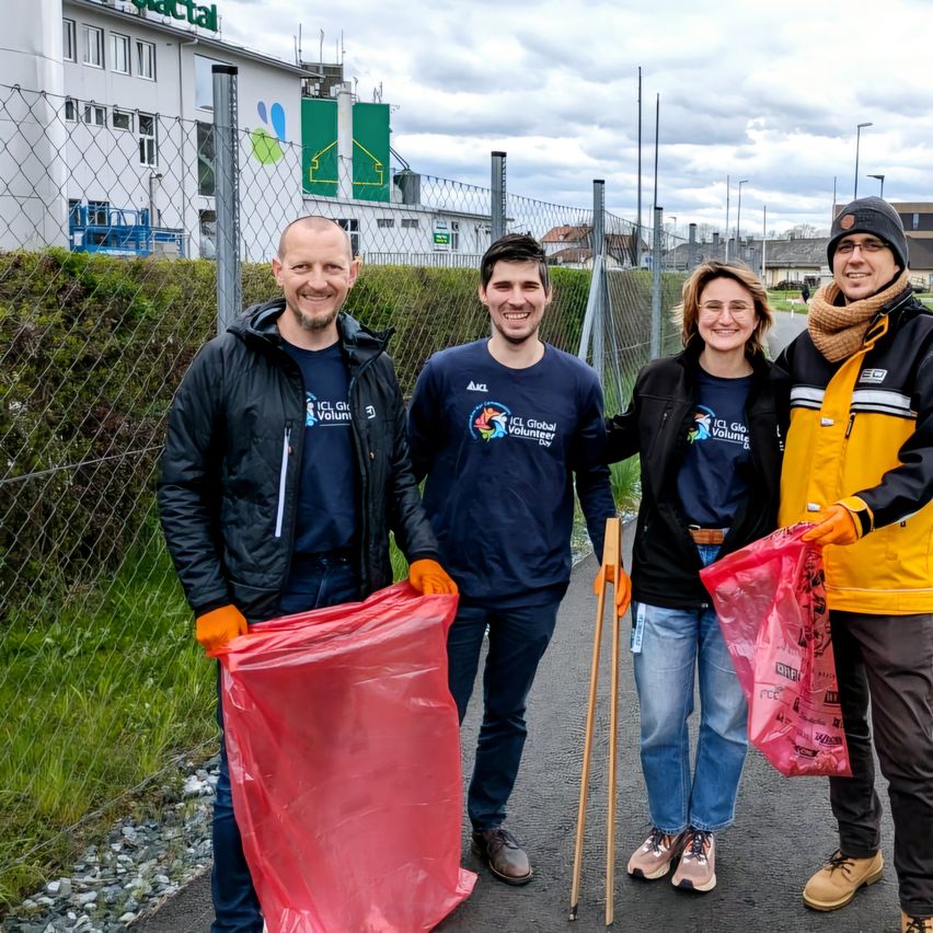 Drei Personen stehen vor einem Zaun mit roten und gelben Taschen in den Händen. Der Mann auf der linken Seite hält eine rote Tasche, während die anderen beiden gelbe Taschen halten. Hinter ihnen steht ein Gebäude mit einem grünen Schild.