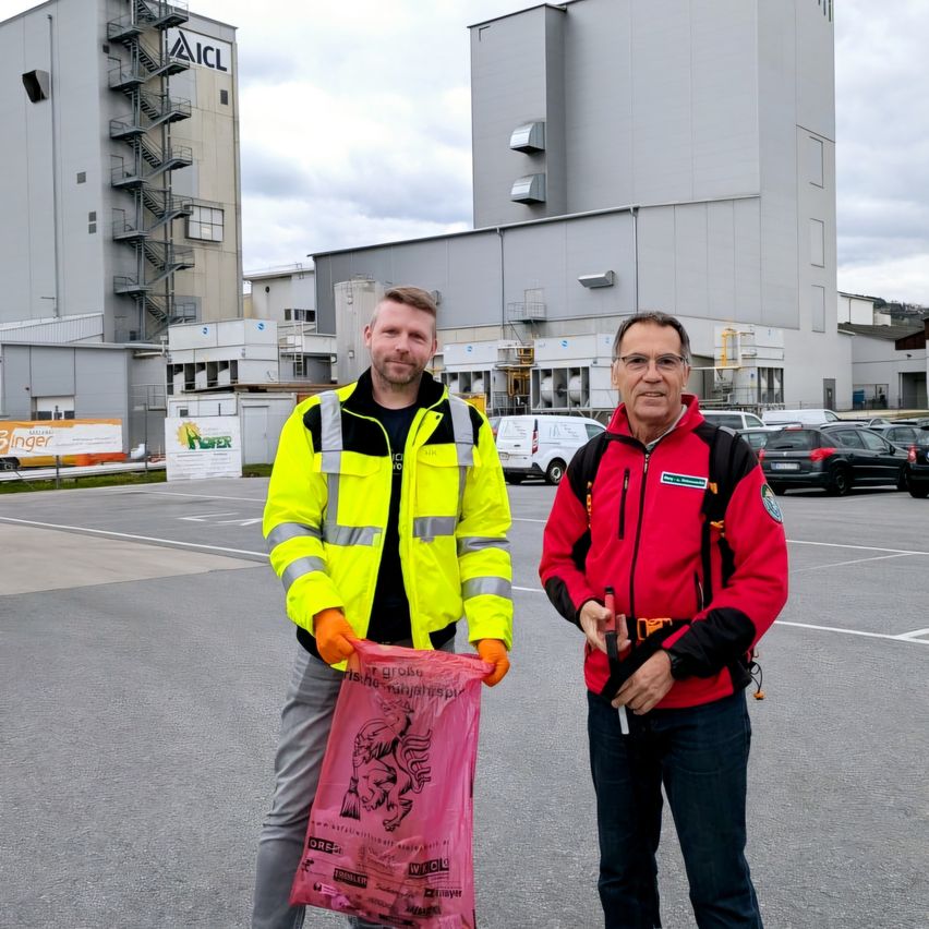Zwei Männer stehen vor einem Gebäude mit einem großen rosa Beutel. Einer von ihnen hält den Beutel und beide tragen orange Handschuhe. Hinter ihnen ist ein großes Gebäude mit dem Namen AIC zu sehen, zusammen mit mehreren geparkten Autos auf einem Parkplatz.