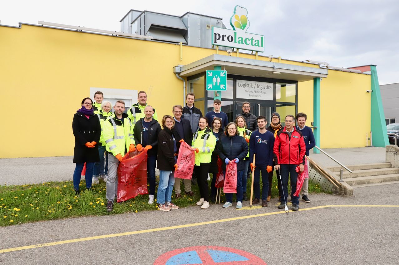 Eine Gruppe von Menschen in Sicherheitswesten und Handschuhen steht vor einem gelben Gebäude mit dem Schild 'prolactal' und einem grünen Schild mit der Aufschrift 'Logistik / logistics'. Sie halten rote Taschen und einige halten Besen. Das Gebäude hat eine Glastür mit der Aufschrift 'An- und Abmeldung'.