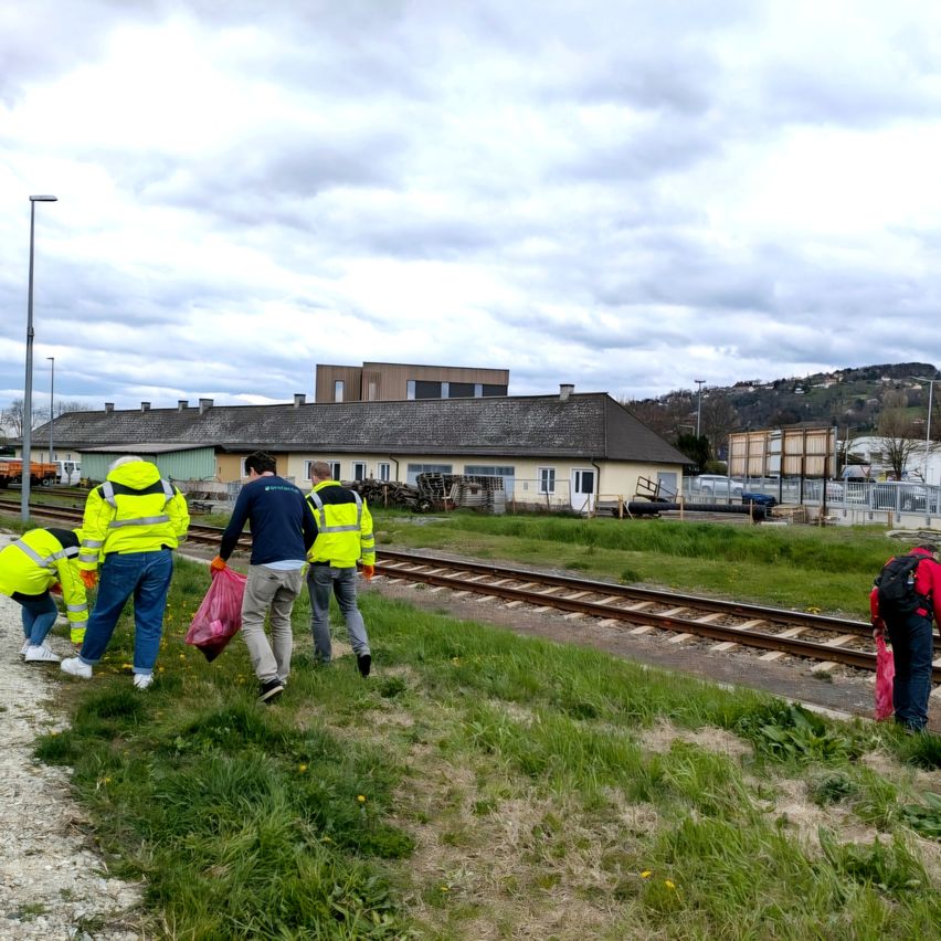 Eine Gruppe von Menschen in reflektierenden gelben Westen geht entlang eines Bahngleises und sammelt Müll. Der Bahndamm ist von Gras umgeben, und im Hintergrund befinden sich Gebäude und Hügel unter einem bewölkten Himmel.