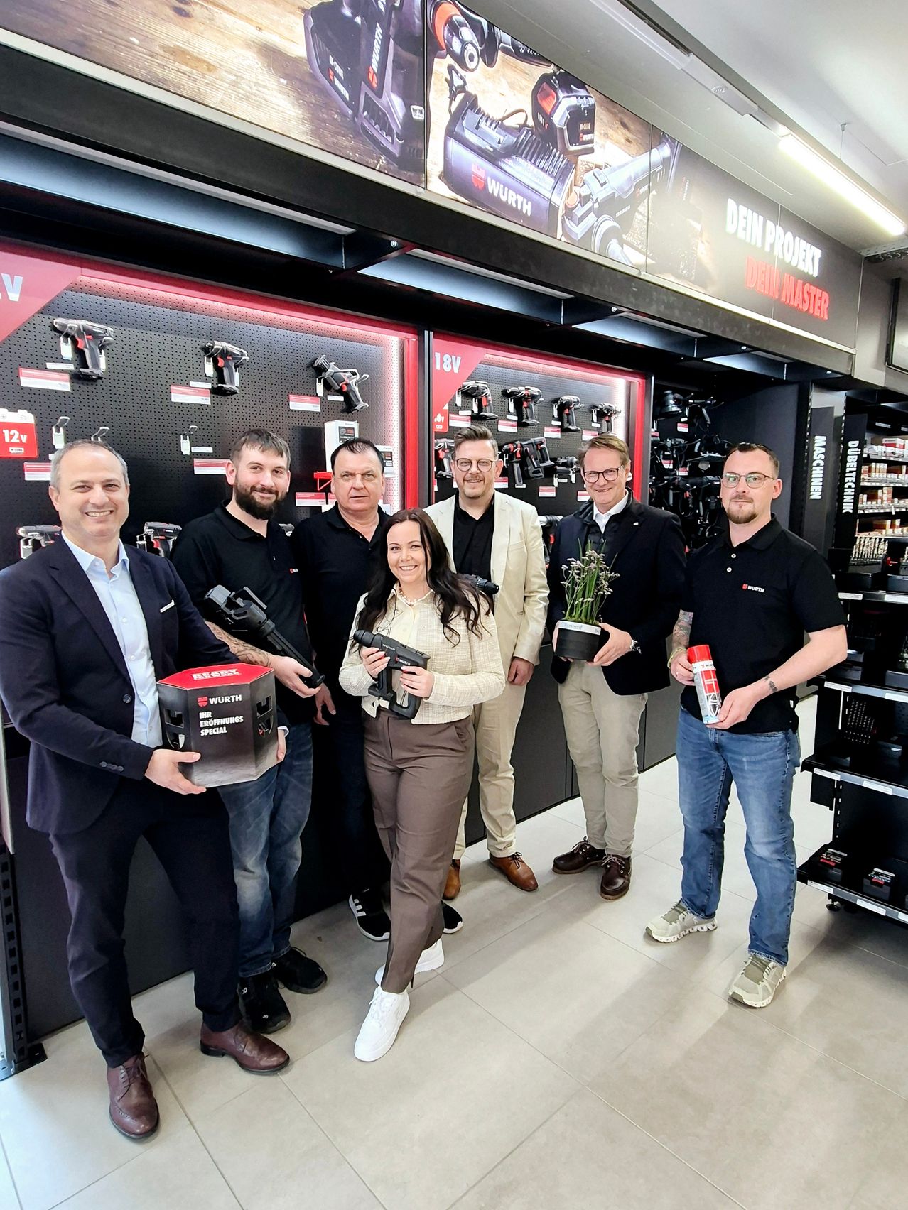 Eine Gruppe von Männern und Frauen steht vor einem Geschäft und hält Werkzeuge und eine Topfpflanze.