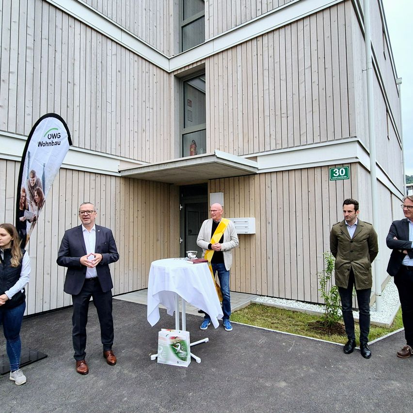 Fünf Personen stehen vor einem Gebäude mit einem Tisch und einem Banner. Der Mann in der Mitte trägt eine gelbe Schärpe.